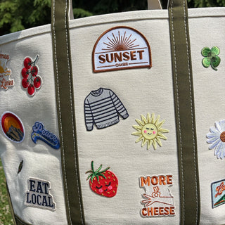 Tote bag with various patches including 'Sunset', 'Eat Local', and 'More Cheese' on a grassy background.