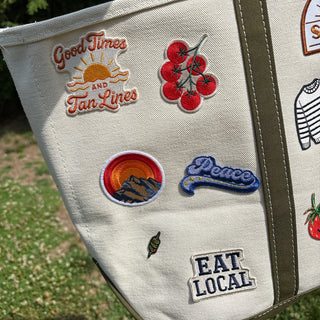 Beige tote bag with various patches including 'Good Times and Fun Lines', 'Peace', and 'Eat Local'.