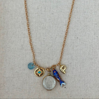 A gold-tone charm necklace laid flat on a light beige fabric background. The necklace has a delicate chain and several charms, including a large oval stone pendant, a blue fish charm, a gold heart charm, a turquoise eye-of-providence charm, and a small light blue crescent charm.