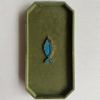 A small fish-shaped jewelry piece with blue and gold detailing, displayed on an olive green velvet jewelry tray over a light beige textured card on a marble surface.