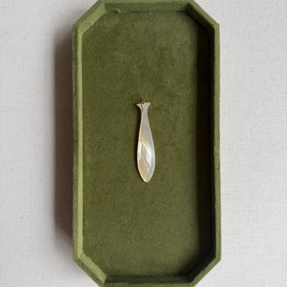 A small mother of pearl fish pendant or charm with a pear-shaped outline and a small floral detail at the top, displayed in the center of an olive green velvet jewelry tray on a light beige fabric surface.