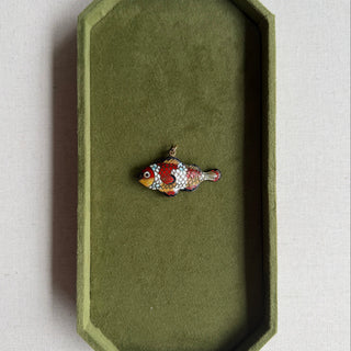 A small fish-shaped enamel charm with red, orange, white, and black detailing rests inside an olive green velvet jewelry box on a light beige fabric surface.