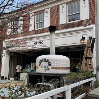 Brick building with 'ohHo' sign and outdoor pizza oven, surrounded by trees and tables.