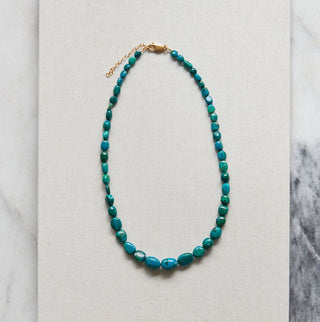 A beaded necklace with turquoise-blue beads and a gold clasp, displayed on a white rectangular card over a white and gray marble surface.