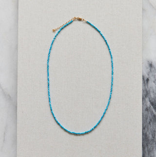 A delicate beaded necklace in bright turquoise blue with a small gold clasp, displayed on a square beige fabric card over a white and gray marble surface.