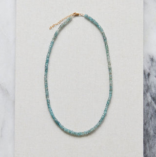 A delicate beaded necklace with small blue-green beads and a rose gold clasp, displayed on a square white card over a gray and white marble surface.