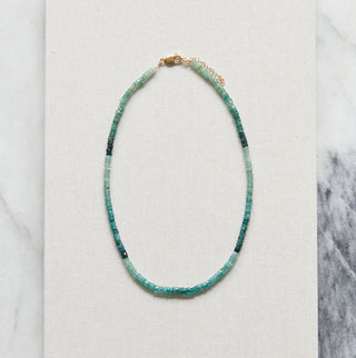 A delicate beaded necklace with turquoise and dark green beads and a small gold clasp, displayed on a white rectangular card over a gray and white marble surface.