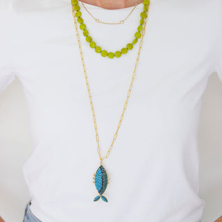 Person wearing a white shirt and blue jeans with layered necklaces, including a green beaded necklace and a gold chain with a blue pendant.
