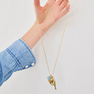 Hand wearing a gold necklace with a blue pendant against a white background