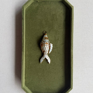 A small fish-shaped jewelry charm with gold and white scales, displayed inside an olive green velvet-lined octagonal tray on a light beige textured surface.