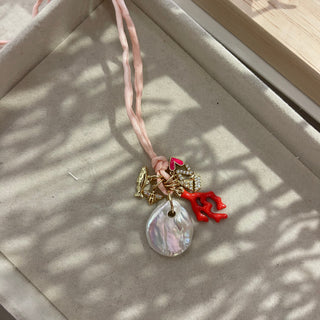 A delicate necklace with a light pink woven cord, a large iridescent shell pendant, and small gold-toned charms including a red coral accent, displayed on a beige jewelry box.
