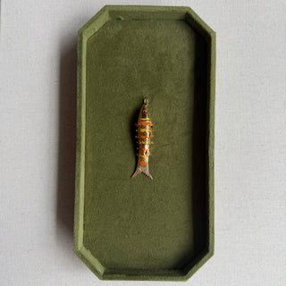 A small decorative fish-shaped pendant or charm with gold, orange, and dark detailing, displayed inside an olive green octagonal tray on a light textured surface.