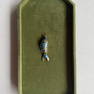 A small fish-shaped jewelry charm with blue, gold, and white enamel details, displayed inside an olive green velvet jewelry box on a light beige fabric surface.