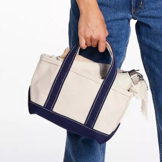 A person wearing blue jeans holds a small tote bag with cream canvas panels and dark navy trim and handles. A silver water bottle is tucked into one of the side pockets, shown against a plain white background.