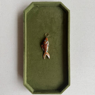 A small fish-shaped pendant with red, orange, and white enamel details and a gold-tone ring at the top, displayed inside an olive green velvet jewelry box on a light beige textured surface.