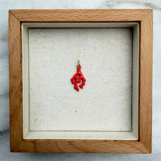 A small red pendant or charm with a gold top loop, displayed inside a square wooden shadow box with a beige fabric lining, set on a gray and white marble surface.