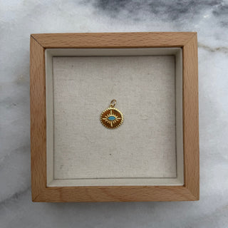 A small round gold pendant with a turquoise blue center, displayed inside a square light wood shadow box on a gray and white marble surface.