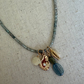 A close-up of a beaded necklace with a gold-toned charm setting featuring a white stone, a textured gold shell-shaped charm with a red center, a small green bead, and a blue faceted bead, displayed on a beige fabric background.